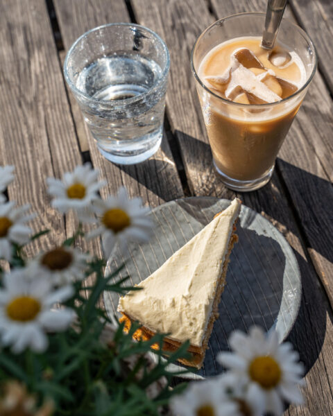 Iskaffe, tårtbit och vattenglas på ett solbelyst träbord med prästkragar, kafé vid Löfstad slott i Norrköping