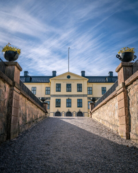 Löfstad slotts gulfärgade huvudbyggnad sedd från grusgången mellan stenmurarna vid entrén, under en klarblå himmel med slöjmoln.