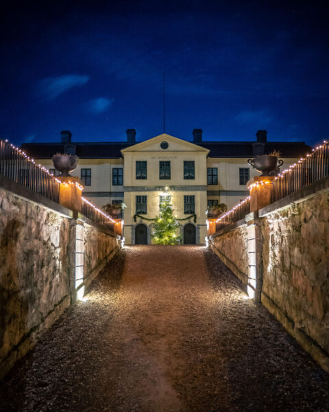 Lövstads slott upplyst i julbelysning en vinterkväll, med ljusslingor längs stenmurarna och en julgran framför entrén.