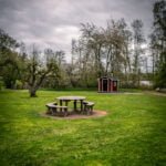 Rastplats vid Torpet Sjöbo i Motala kommun med runt träbord på grön gräsmatta, litet rött dass och blommande fruktträd under mulen himmel.
