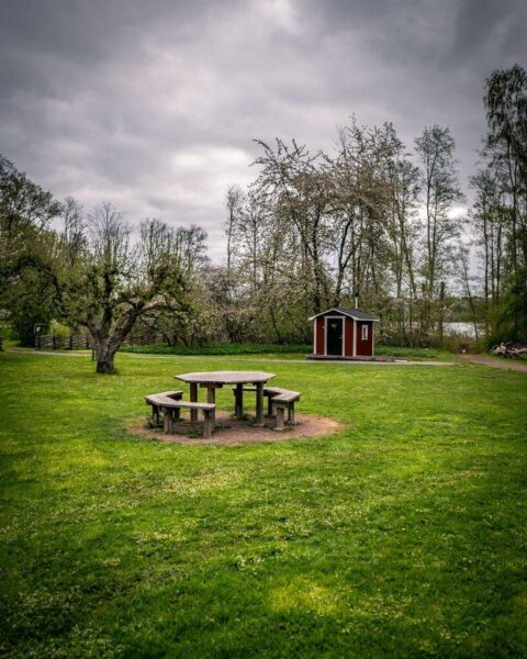 Rastplats vid Torpet Sjöbo i Motala kommun med runt träbord på grön gräsmatta, litet rött dass och blommande fruktträd under mulen himmel.