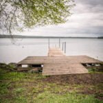 Träbrygga vid Knäppaviken i Motala med utsikt över stilla vatten en mulen vårdag, omgiven av grönt gräs och lövträd.