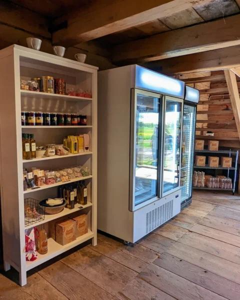 Gårdsbutik med lokala produkter i rustik timmerbyggnad, hylla med sylt, oljor och delikatesser samt ett kyldiskskåp.