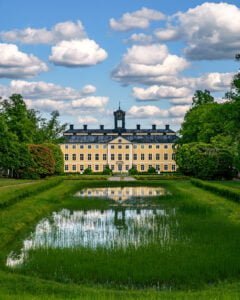 Gult barockslott med klocktorn speglas i en damm omgiven av gröna gräsmattor och lummiga träd en sommardag med vitblå himmel.
