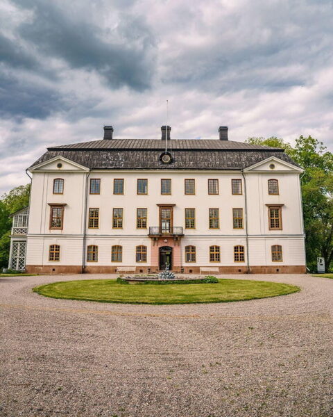 Ljungs slotts vitputsade huvudbyggnad med grusplan, grön gräsmatta och fontän framför entrén under dramatisk molnhimmel.