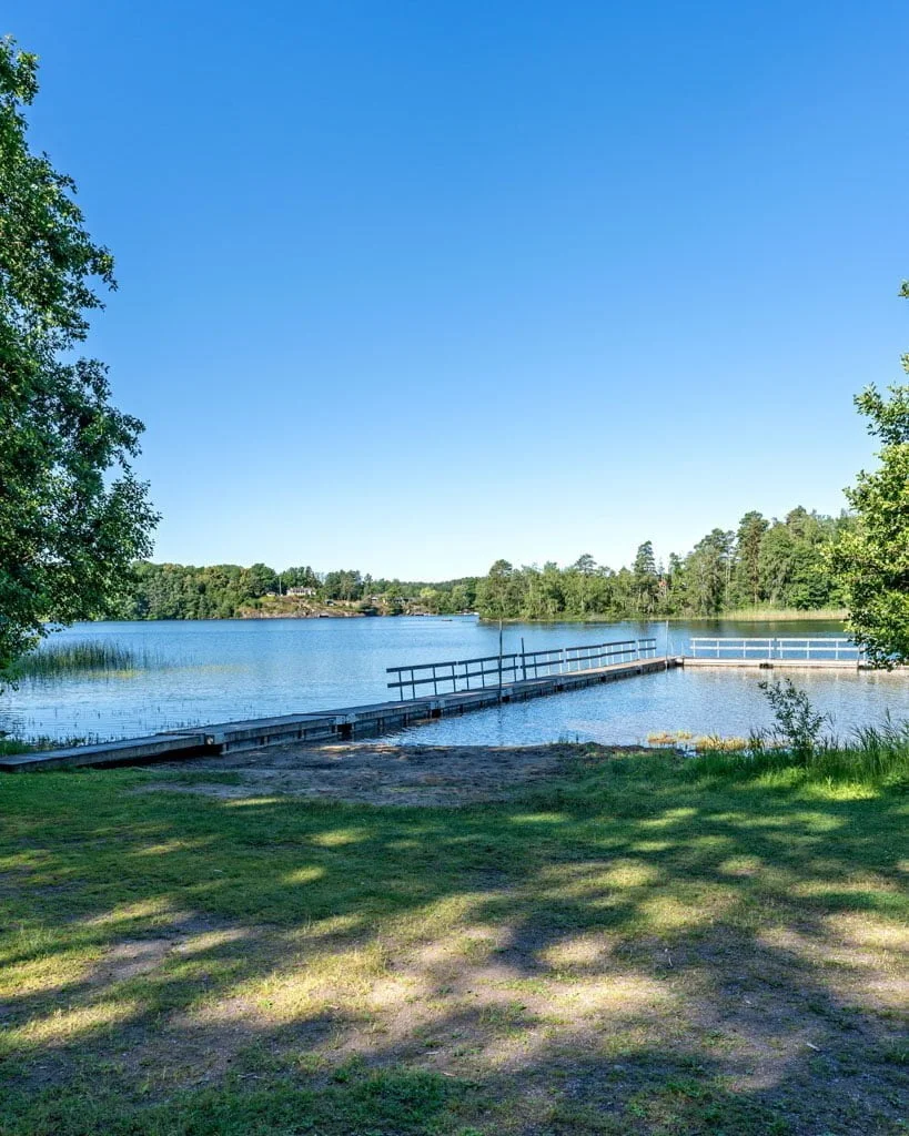 Badbrygga vid Viggebysandsbadet med stilla vatten, gröna gräsytor och lummig skog i bakgrunden en solig sommardag.