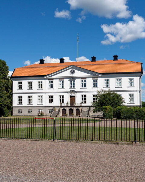 Brokinds herrgård med vit fasad, orangerött tegeltak och flaggstång, omgärdad av svart järnstaket och grön gräsmatta en solig sommardag.