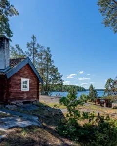 Röd timmerstuga på Jaktstugeudden vid Viggeby naturreservat med klippor, tallar och sjöutsikt en solig sommardag.