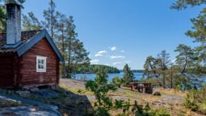 Röd timmerstuga med skorsten vid Jaktstugeudden i Viggeby naturreservat, med utsikt över sjön, klippor, tallar och picknickbord.
