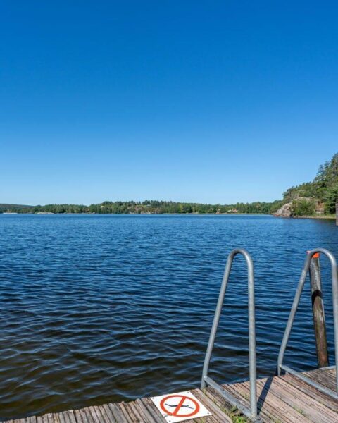Brygga med badstege vid Jaktstugeuddens badplats i Viggeby naturreservat, med utsikt över stilla vatten och skogsklädd strand en solig sommardag.
