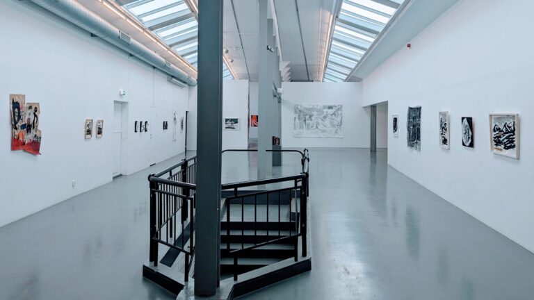 Ljus konsthallssal på Passagen i Linköping med konstverk på vita väggar, svart spiraltrappa och takfönster.