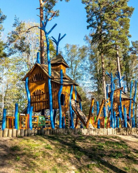 Fantasifullt lekhus i trä omgivet av blå dekorativa trädstammar på Lill-Valla Aktivitetspark i Linköping, med tallskog i bakgrunden
