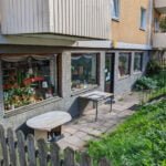 Liten butik i gatuplan vid flerfamiljshus i Linköping med skyltfönster fyllda av växter och prylar, omgärdat av trästaket och grönska