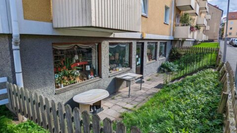Liten butik i gatuplan vid flerfamiljshus i Linköping med skyltfönster fyllda av växter och prylar, omgärdat av trästaket och grönska