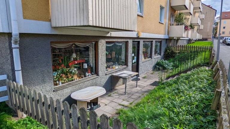 Liten butik i gatuplan vid flerfamiljshus i Linköping med skyltfönster fyllda av växter och prylar, omgärdat av trästaket och grönska