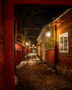 Smal kullerstensgränd i Gamla Linköping en vinterkväll, med röda och gula trähus, lyktor och lätt snö på marken.