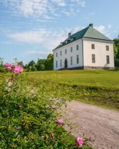 Verner von Heidenstams vitputsade herrgård Övralid med grönt tak, omgiven av gröna gräsmattor och rosa rosor en sommardag