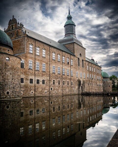Vadstena slott med tegelfasad och koppartak speglar sig i den stilla vallgraven under dramatisk molnhimmel