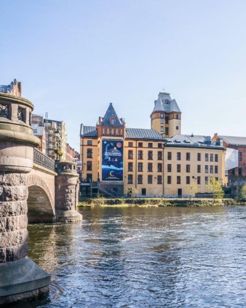 Visualiseringscenter C vid Motala ström i Norrköpings industrilandskap, med historiska tegelbyggnader och en stenbro en solig dag