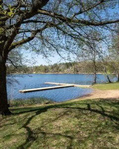 Badbrygga i solsken vid Hultsjöns badplats nära Ljungsbro, med grön gräsmatta, lövträd och stilla vatten omgivet av skog.