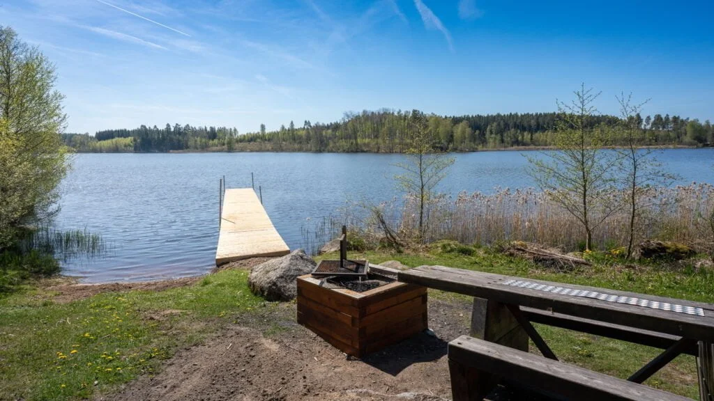 Badbrygga vid Sibborpesjöns badplats utanför Ljungsbro med grillplats, bänk och stilla sjövatten omgivet av skog en solig vårdag