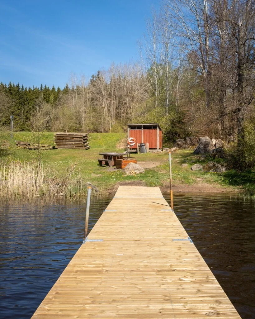 Ny badbrygga i trä vid Sibborpesjöns badplats norr om Ljungsbro, med röd omklädningshytt, livboj och picknickbord på gröna gräset.