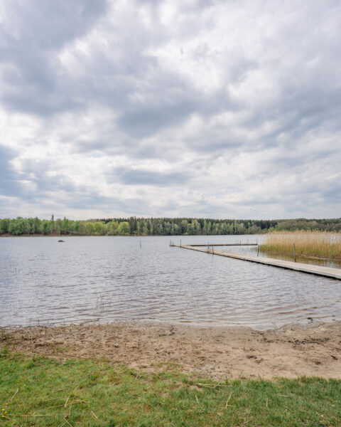 Lillsjöns badplats i Gammalkil med sandstrand, lång badbrygga och stilla vatten omgivet av skog under molnig himmel.