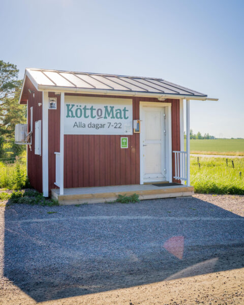 Liten faluröd självservicebutik med skylten "KöttoMat – Alla dagar 7-22" vid Fröö Gård, omgiven av gröna åkrar i Östergötland.