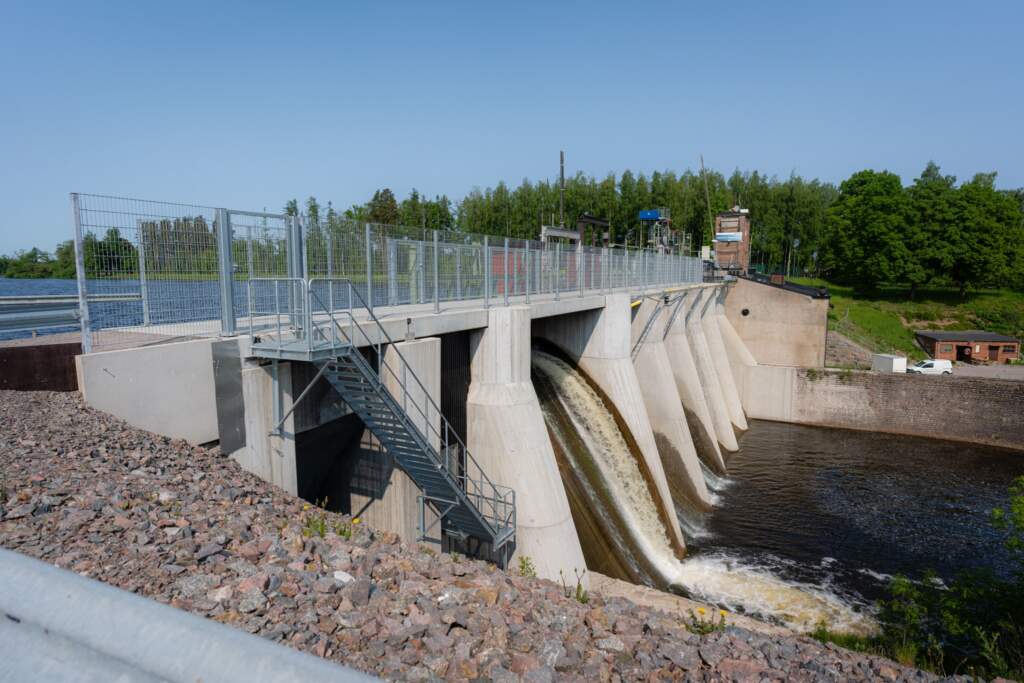 Betongdamm vid Svartåfors med vatten som forsar genom utskovsluckorna, omgiven av skog och stenbelagd sluttning en solig dag.