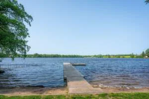 Träbrygga vid Svartåfors badplats som sträcker sig ut i en stilla sjö, omgiven av grönska och blå himmel en sommardag.