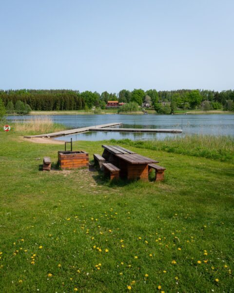 Grillplats och sittbänk i trä vid Ljungbadet med badbrygga, stilla sjö och röda stugor omgivna av grönska en solig sommardag.