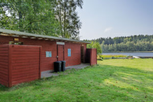 Röd servicebyggnad med omklädningsrum och toaletter vid Amundebobadet i Ulrika, med grön gräsmatta och sjön i bakgrunden