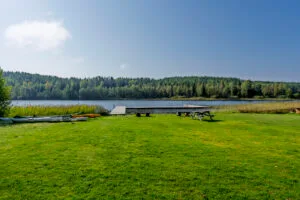 Grön gräsmatta vid Amundebobadet i Ulrika med kanoter, brygga, picknickmöbler och utsikt över sjön omgiven av skog.