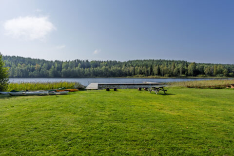 Grön gräsmatta vid Amundebobadet i Ulrika med kanoter, brygga, picknickmöbler och utsikt över sjön omgiven av skog.