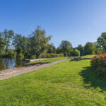 Johannelundsbadet vid Stångån i Linköping med gröna gräsytor, picknickbord, badlagun och lummiga träd under klarblå himmel.