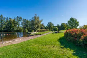 Johannelundsbadet vid Stångån i Linköping med gröna gräsytor, picknickbord, badlagun och lummiga träd under klarblå himmel.