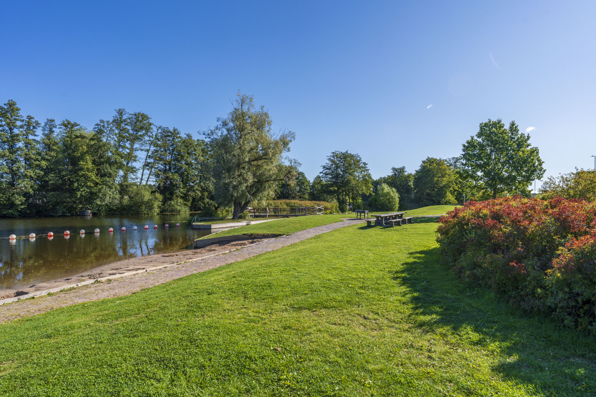 Johannelundsbadet vid Stångån i Linköping med gröna gräsytor, picknickbord, badlagun och lummiga träd under klarblå himmel.