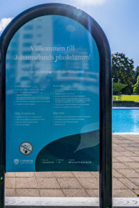 Informationsskylt för Johannelunds plaskdamm i Linköping med ordningsregler, pool synlig i bakgrunden.