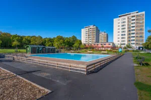 Johannelunds plaskdamm i Linköping med träkantad pool, grönskande träd och höghus i bakgrunden under klarblå himmel.