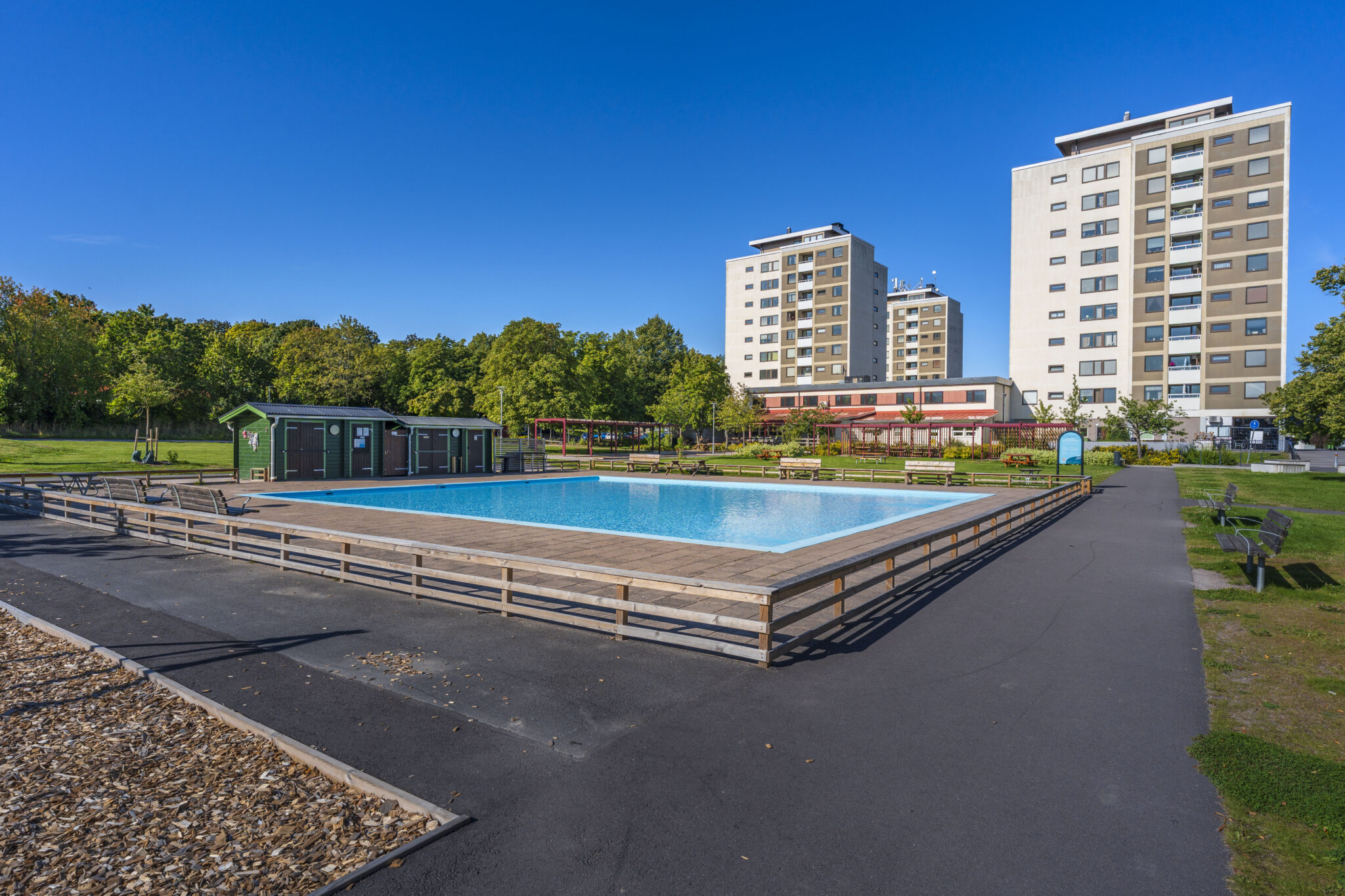 Johannelunds plaskdamm i Linköping med träkantad pool, grönskande träd och höghus i bakgrunden under klarblå himmel.