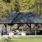 Grillplats med tak, bänkar och grillringar vid Lill-Valla aktivitetspark i Linköping, omgiven av grönska en solig vårdag