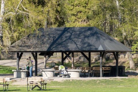 Grillplats med tak, bänkar och grillringar vid Lill-Valla aktivitetspark i Linköping, omgiven av grönska en solig vårdag