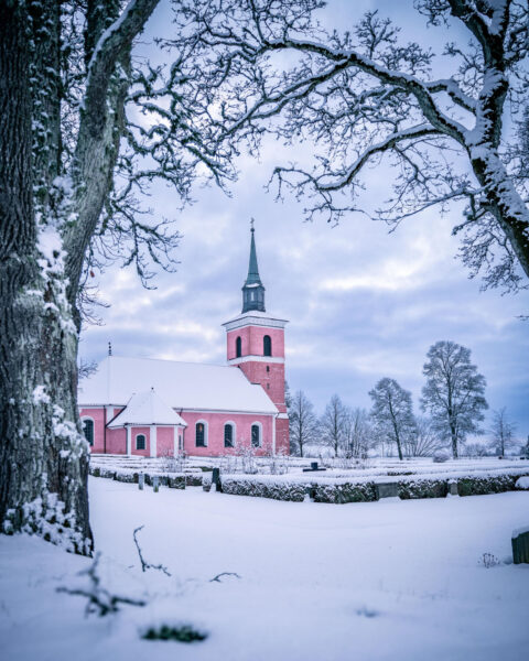 Slaka kyrka med rosa puts omgiven av snötäckt kyrkogård och frostiga träd en vinterdag i Östergötland