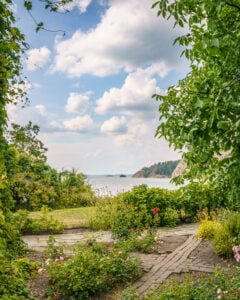 Lummig trädgård med rosor och stengång vid Stegeborgs slottsruin, med utsikt över Slätbaken och skogklädda klippor en sommardag