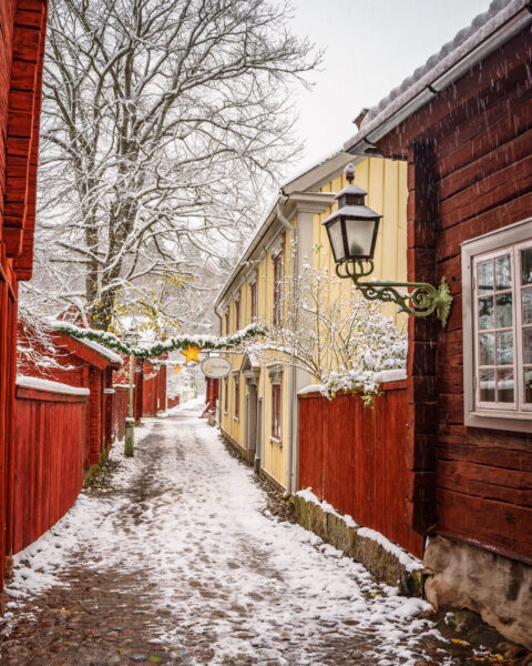 Snötäckt gränd i Gamla Linköping med faluröda trähus, gult hus, lykta och juldekorationer med stjärna och granrisgirlang.