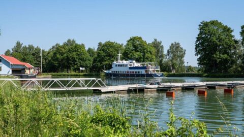Passagerarfartyget Wasa Lejon vid bryggan i Malfors hamn, omgivet av grönskande träd och lugnt vatten en solig sommardag.