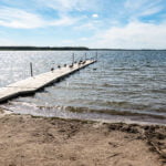 Lång träbrygga med fåglar sträcker sig ut i glittrande vatten vid Strandbadet i Borensberg, sandstrand i förgrunden