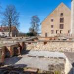 En solig dag vid Vadstena klosterruin. I förgrunden syns de bevarade stenfundamenten från klostrets byggnader, med tegelvalv och murade strukturer. Vadstena klosterkyrka ligger intill. Bakom detta hus står en större byggnad med beige puts och röda fönsterkarmar. Höga träd ramar in scenen, och i bakgrunden skymtar en glimt av ett blått vattenlandskap.