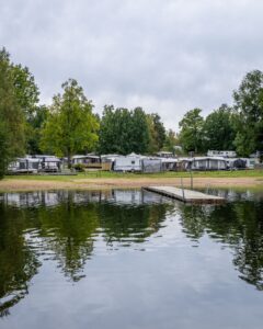 Småängsbadet i Motala med badbrygga, stilla vatten och sandstrand framför en campingplats med husvagnar omgivna av gröna lövträd.