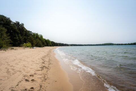 Varamobadets långa sandstrand vid Vättern i Motala, med mjuk sand, små vågor och tallskog längs strandkanten.